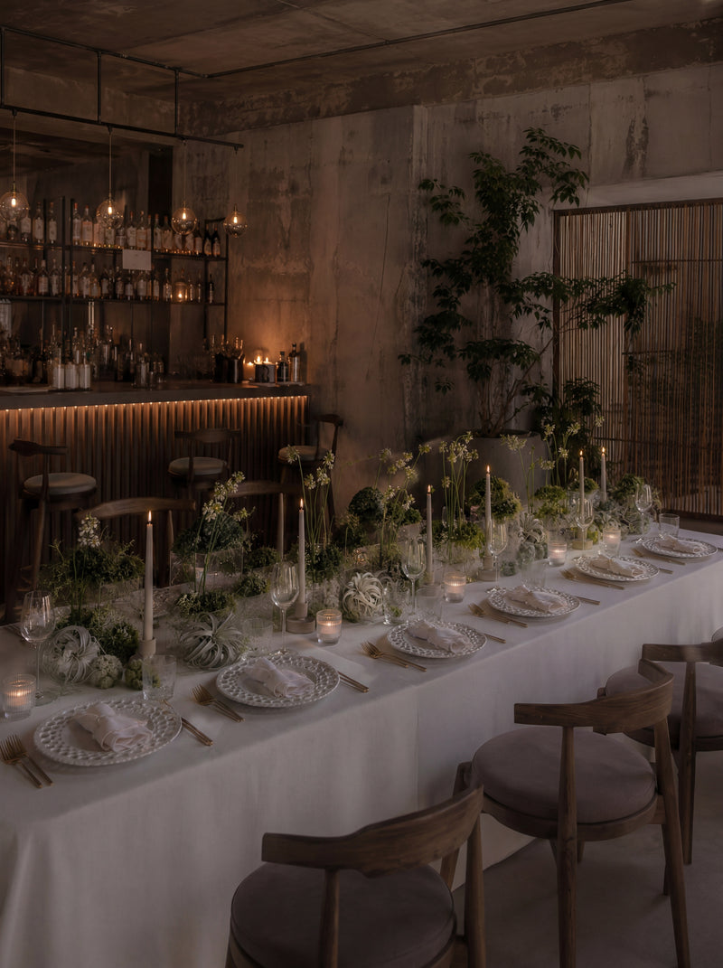 Long dining table with sculptural floral arrangements, candles, and modern tablescape design in a moody restaurant setting.